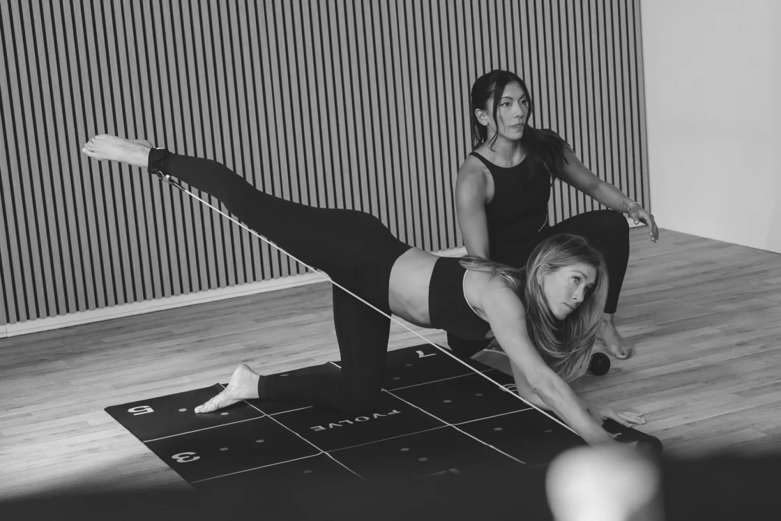 Woman performs a resistance-based lower-body exercise on a Pvolve mat while a Pvolve trainer assists with form in a minimalist studio.