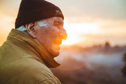 Elderly man in a beanie and jacket gazes toward the sunrise, his face lit by warm morning light against a softly blurred landscape.