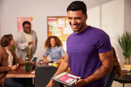 Smiling employee holding a tablet with a wellness app open while colleagues chat in a modern office setting.